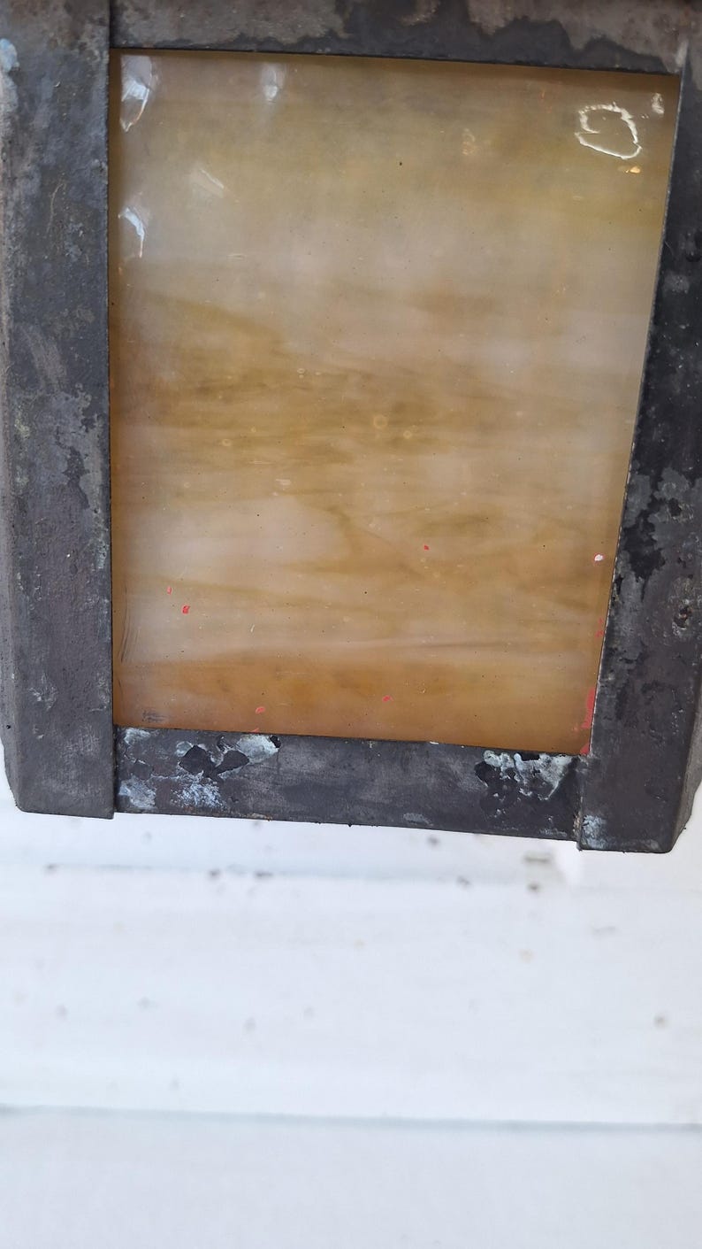 May include: A rectangular wall-mounted lantern featuring a weathered, dark metal frame with visible signs of oxidation and wear. The central panel consists of a translucent, amber-toned glass with subtle swirling patterns and small red paint flecks.