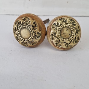 May include: Two antique door knobs with a wooden base and ornate brass detailing. The brass features a circular design with a beaded center and swirling patterns around the edge. The knobs are set against a white background.
