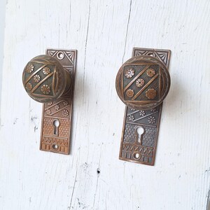 May include: Two antique brass door knobs with intricate floral designs and keyholes. The knobs are mounted on rectangular plates with matching designs.