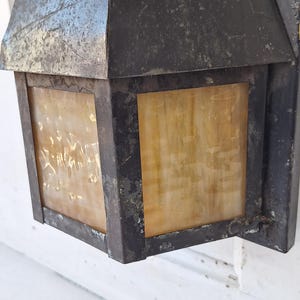 May include: A wall-mounted outdoor lantern featuring a dark, weathered metal frame with a distressed finish. The fixture includes rectangular panels of translucent, amber-toned glass with a mottled, textured appearance and a peaked metal roof.