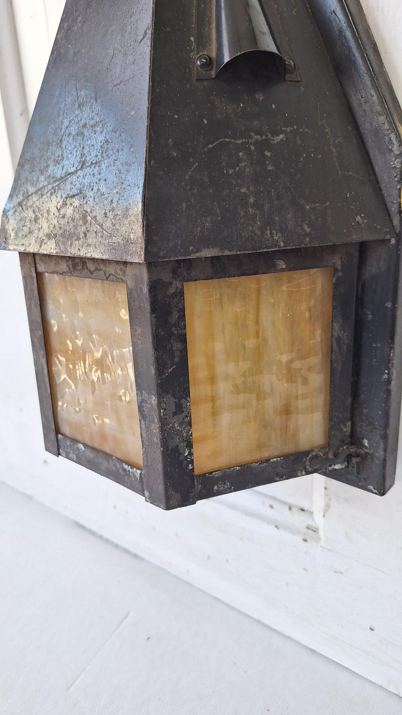 May include: A wall-mounted outdoor lantern featuring a dark, weathered metal frame with a distressed finish. The fixture includes rectangular panels of translucent, amber-toned glass with a mottled, textured appearance and a peaked metal roof.