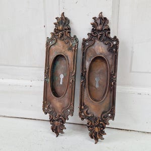 May include: A pair of antique ornate brass door plates with keyhole cutouts. The plates have a distressed finish and intricate floral designs.