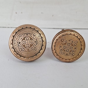May include: Two antique bronze-colored cabinet knobs. Each knob has a circular design with an embossed floral pattern in the center, surrounded by a detailed border. The knobs are likely made of metal and are designed for furniture.