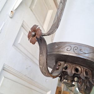 K&ouml;nnte beinhalten: Nahaufnahme einer antiken Deckenleuchte mit wei&szlig;em Glasschirm. Der Metallrahmen hat ein abgenutztes, gealtertes Aussehen mit floralen und scrollartigen Details. Die Leuchte h&auml;ngt in der N&auml;he einer wei&szlig;en T&uuml;r.