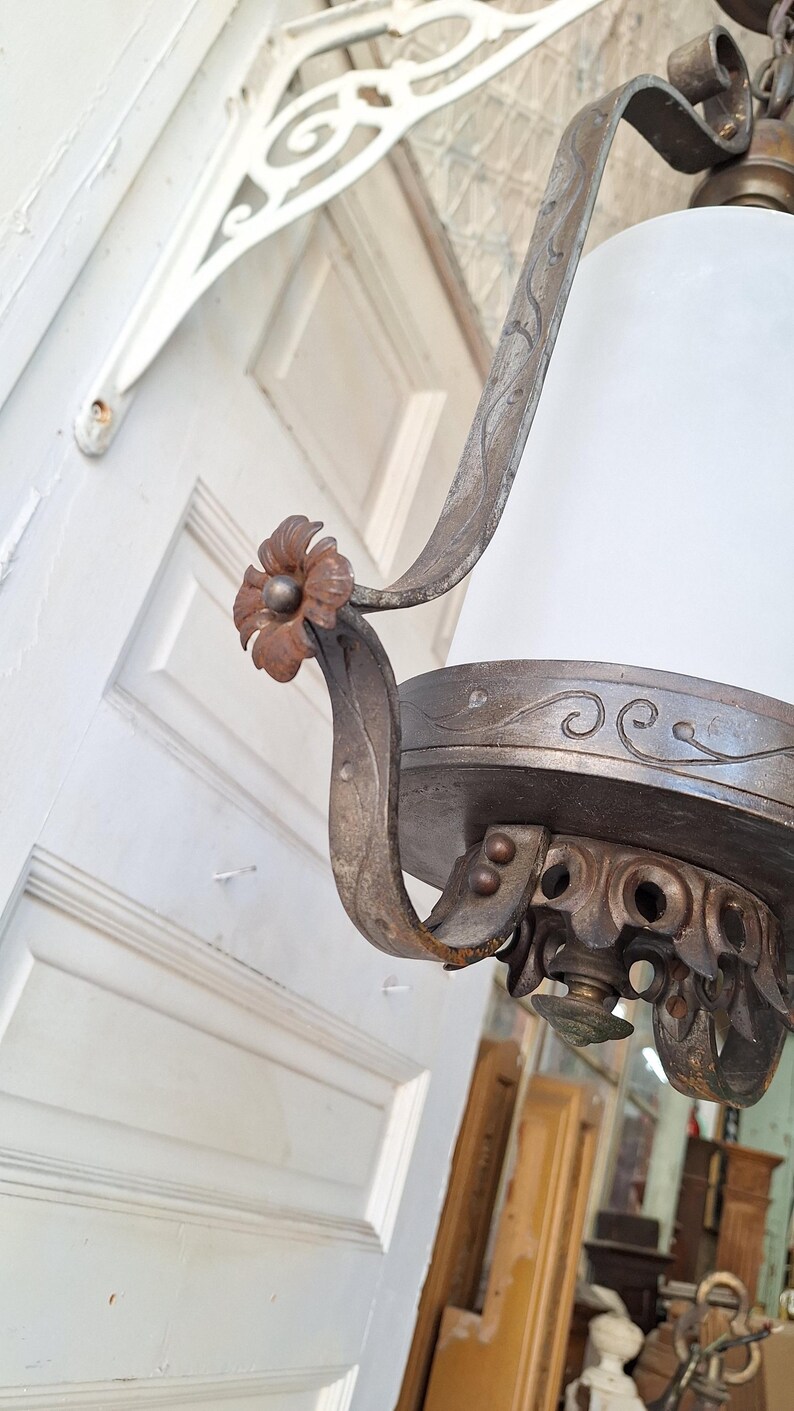 K&ouml;nnte beinhalten: Nahaufnahme einer antiken Deckenleuchte mit wei&szlig;em Glasschirm. Der Metallrahmen hat ein abgenutztes, gealtertes Aussehen mit floralen und scrollartigen Details. Die Leuchte h&auml;ngt in der N&auml;he einer wei&szlig;en T&uuml;r.