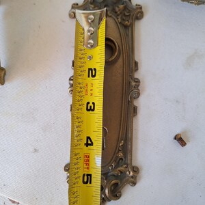 Puede incluir: Herrajes de puerta antiguos de lat&oacute;n ornamentado, que incluyen una placa trasera decorativa de aproximadamente 14 cm de largo. Los herrajes presentan intrincados adornos y un ojo de cerradura. Una cinta m&eacute;trica se coloca junto a los herrajes.
