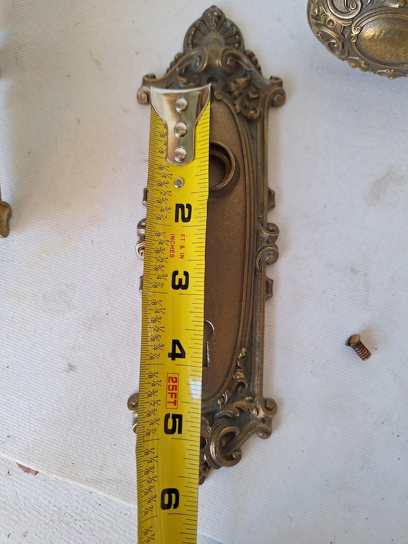 Puede incluir: Herrajes de puerta antiguos de lat&oacute;n ornamentado, que incluyen una placa trasera decorativa de aproximadamente 14 cm de largo. Los herrajes presentan intrincados adornos y un ojo de cerradura. Una cinta m&eacute;trica se coloca junto a los herrajes.
