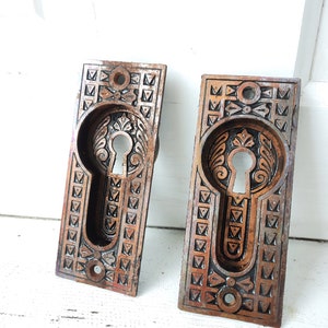 May include: Two antique keyhole escutcheons with ornate designs in a copper finish. The escutcheons are rectangular with a keyhole in the center and intricate details around the edges.