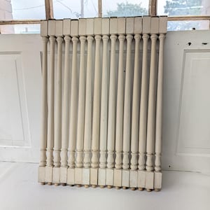 May include: A set of twelve off-white wooden balusters arranged in a vertical row. Each baluster features a square base, a turned spindle section with a rounded profile, and a square top block, all finished in a uniform matte off-white paint.