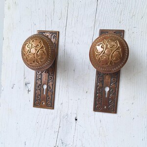 May include: Two antique brass door knobs with ornate floral designs and keyholes. The knobs are mounted on rectangular plates with matching designs.