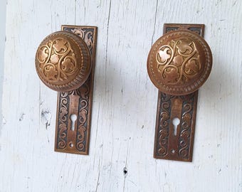 May include: Two antique brass door knobs with ornate floral designs and keyholes. The knobs are mounted on rectangular plates with matching designs.