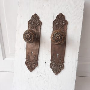 May include: A pair of antique brass door knobs with ornate designs. The knobs are mounted on a white wooden surface.
