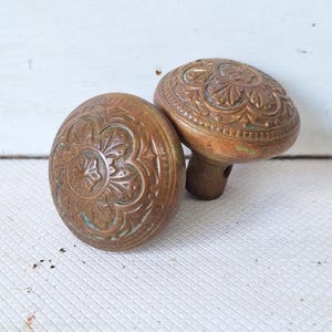 May include: Two antique bronze door knobs with intricate floral and scrollwork designs. The knobs have a warm, aged patina, and are set against a white background. The knobs are oval-shaped and have a stem for mounting.