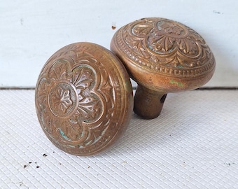 May include: Two antique bronze door knobs with detailed floral and scrollwork patterns. The knobs have a warm, aged patina, set against a white background. The knobs are oval-shaped and have a stem for mounting.
