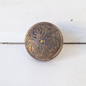 May include: A round, antique brass button with an ornate, symmetrical floral design. The button features a central gold-colored dot and detailed, swirling patterns. The button is set against a white, textured background.