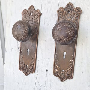 May include: Two antique bronze door knobs with ornate floral detailing. Each knob is round and mounted on a rectangular backplate with a keyhole. The hardware is set against a weathered, white-painted wooden surface.
