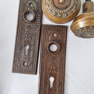May include: Antique bronze door hardware set, including two rectangular backplates with keyholes and decorative patterns, and two round doorknobs. The hardware has a vintage, aged appearance.