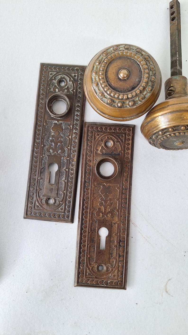May include: Antique bronze door hardware set, including two rectangular backplates with keyholes and decorative patterns, and two round doorknobs. The hardware has a vintage, aged appearance.