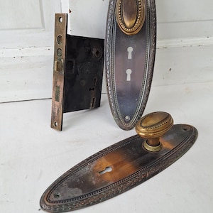 May include: Antique door hardware set featuring two oval backplates with keyholes and decorative trim, and a brass knob. A separate rectangular lock mechanism is also included. The hardware has a tarnished, aged appearance.
