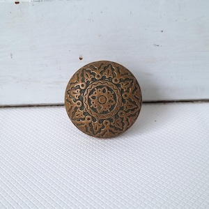 May include: A round, antique bronze knob with an intricate floral and geometric pattern. The knob has a central circular design surrounded by detailed leaf-like shapes. The aged metal has a dark patina.