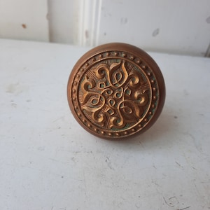 May include: A round, brass door knob with a decorative, ornate design in the center. The design is gold and features a floral pattern. The knob has a beaded edge.