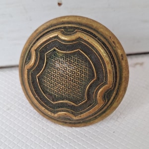 May include: A vintage brass door knob with a decorative, geometric pattern. The knob has a dark patina and a raised, rounded edge.