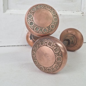 May include: Close-up of antique copper door knobs. The knobs have a circular design with an ornate pattern around the center. The copper has a warm, aged patina, suggesting vintage or reclaimed hardware. The knobs are likely for interior doors.