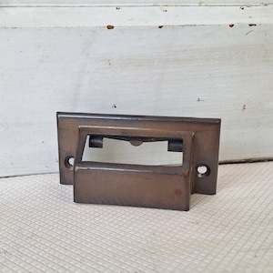 May include: A bronze-colored drawer pull with a rectangular backplate and a curved handle. The pull has two screw holes for mounting. The hardware is likely vintage or antique, suitable for cabinets, drawers, or furniture restoration projects.