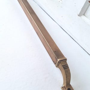 May include: A long, rectangular bronze-colored door pull with a curved end and decorative detailing. The hardware is made of metal and is designed for a door. The door pull is shown against a white background.