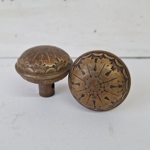 May include: Two antique brass door knobs with intricate floral designs. The knobs have a warm, aged patina, and are likely made of solid brass. One knob is upright, showing the stem, while the other lies on its side.