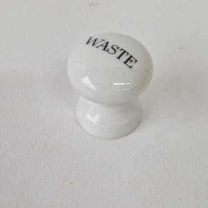 May include: White ceramic knob with the word "WASTE" written in black lettering.