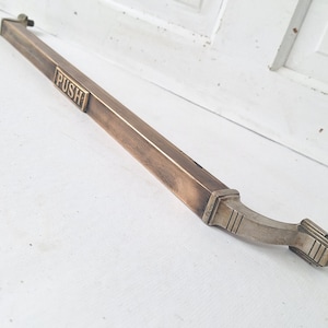 May include: A bronze-toned door pull with the word "PUSH" embossed on the rectangular bar. The handle curves slightly at the end, with a decorative design. The door pull is set against a white background.