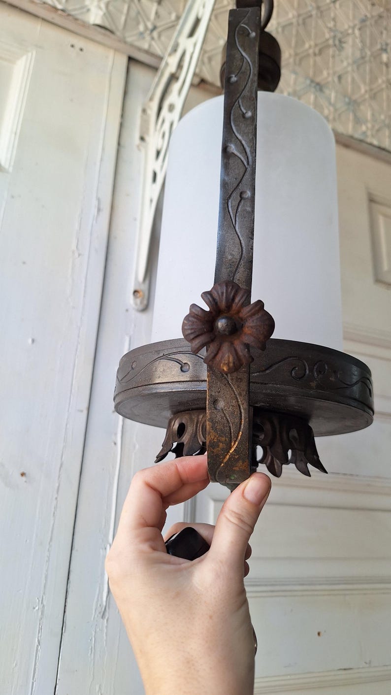 K&ouml;nnte beinhalten: Antike H&auml;ngeleuchte mit einem Schirm aus Milchglas. Die Leuchte hat einen dunklen Metallrahmen mit floralen und Ranken Details. Der handgehaltene Teil ist dunkel gef&auml;rbt. Die Leuchte ist auf einer wei&szlig;en Oberfl&auml;che montiert.