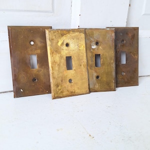 May include: Four antique brass light switch plates with a single toggle switch. The plates are slightly worn and have a patina finish.