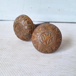 May include: Two antique bronze door knobs with intricate embossed designs. The knobs are round with a central motif surrounded by a detailed border. The bronze has a warm, aged patina, suggesting vintage craftsmanship. The knobs are likely for a door.