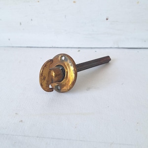 May include: A vintage brass door thumb turn latch featuring a circular mounting plate with two screw holes and a central rotating handle. The hardware includes a long, thin, square-shaped metal spindle extending from the back of the circular plate.