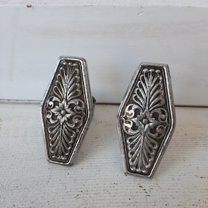 May include: Two silver-toned metal drawer pulls with an ornate floral design. The pulls are shaped like a coffin and have a hexagonal base.