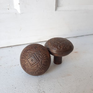 May include: Two antique brass door knobs with intricate floral and geometric designs. The knobs are round and have a raised center.