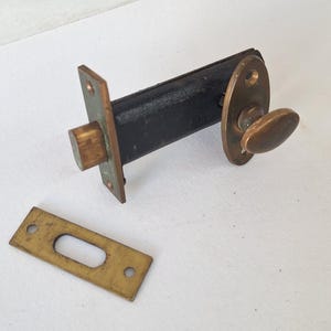 May include: A brass door latch mechanism featuring a rectangular faceplate, a black cylindrical body, and an oval-shaped thumb turn knob. A separate rectangular brass strike plate with two screw holes and a central opening is positioned nearby.