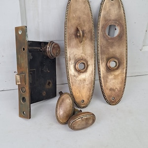 May include: Antique brass door hardware set including a mortise lock, two oval backplates, and three matching oval doorknobs. The hardware has a detailed beaded edge design and shows signs of age.