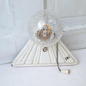 May include: A vintage-style wall sconce featuring a clear, textured globe light bulb. The bulb sits atop a white, triangular ceramic base with a pull chain. The sconce has a classic, retro design, perfect for adding a touch of elegance to any room.