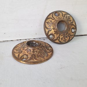 May include: Two antique brass door rosettes with ornate floral designs. The rosettes are circular with a central hole and several smaller holes around the perimeter. The brass has a warm, aged patina, suggesting vintage hardware.