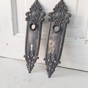 May include: Two antique, ornate door plates with keyholes and circular openings for the doorknob. The plates are dark gray with decorative scrollwork and a distressed finish, suggesting an aged appearance. The door plates are likely vintage or reproduction.