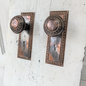 May include: Two matching copper-toned metal door knobs with circular, ribbed handles and rectangular backplates featuring a decorative border and a central keyhole. The hardware displays a weathered, tarnished finish with visible dark patina.