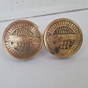 May include: Two antique brass cabinet knobs with a circular design. Each knob features an intricate pattern of lines, circles, and a stylized leaf or fern motif. The knobs have a warm, aged patina, suggesting they are vintage or vintage-inspired.