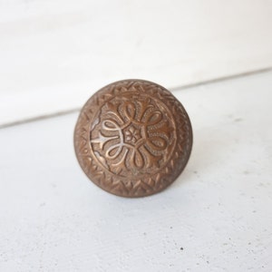 May include: A vintage brass door knob with an intricate, ornate design. The knob is round and has a raised, circular pattern in the center.