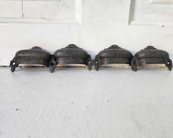 May include: Four antique-style black metal drawer pulls with ornate designs. Each pull has a curved shape and decorative details.