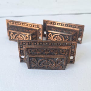 May include: Three rectangular metal drawer pulls with a dark copper finish. Each pull features a recessed center panel with an embossed fan and scroll motif, a decorative border of small squares, and two circular screw holes on the side flanges.
