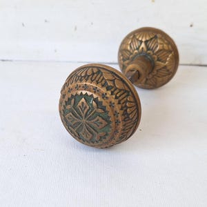May include: Two antique brass door knobs with detailed floral and geometric patterns. The knobs have a weathered, aged appearance, suggesting they are vintage or reclaimed. The design includes a central cross-like motif.