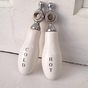May include: Two vintage faucet handles. The handles are white with the words "COLD" and "HOT" in black lettering. The top of each handle is chrome. The handles are likely for a bathroom or kitchen sink.